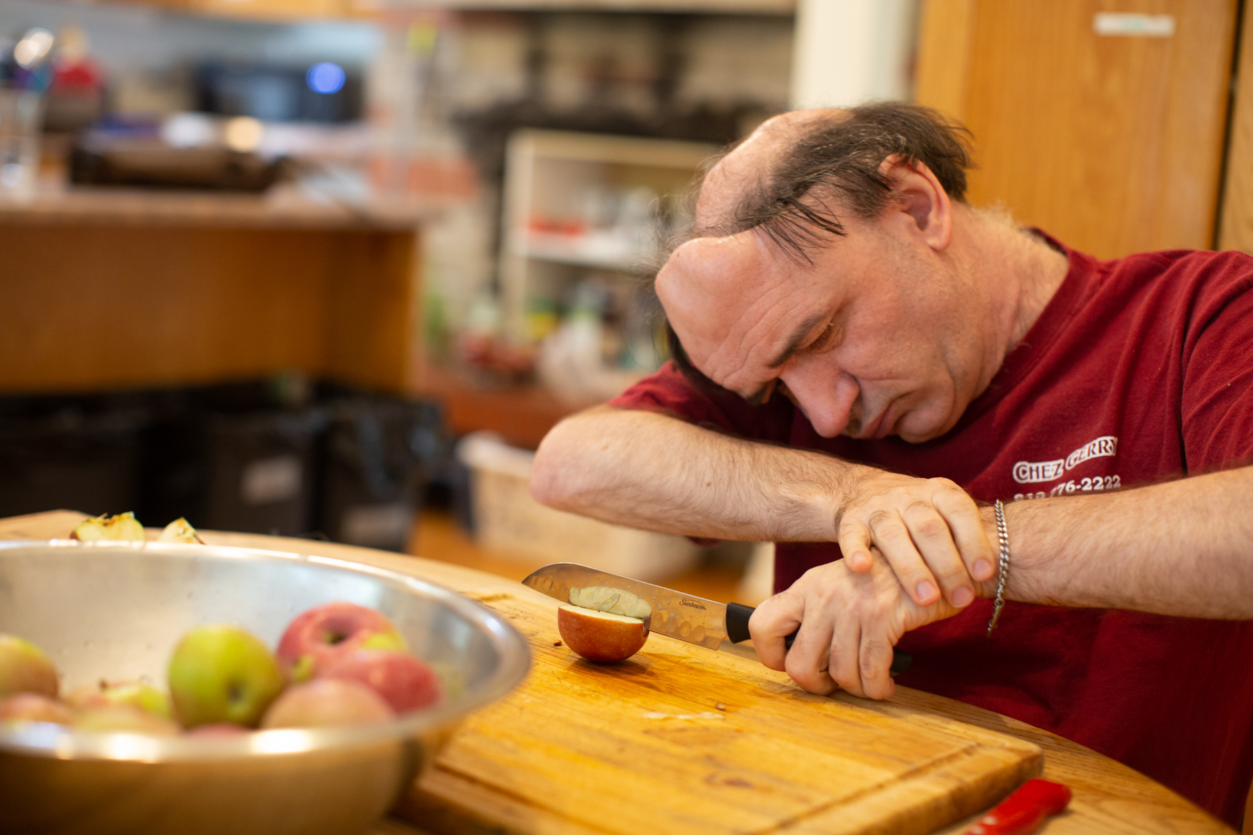 A core member wearing a red t-shirt sits at a table cutting a red apple on a wooden cutting board. A bowl of apples sits in front of the core member.